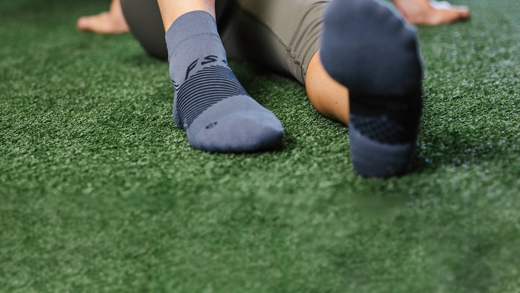 Woman wearing grey plantar fasciitis socks while sitting on a turf floor 